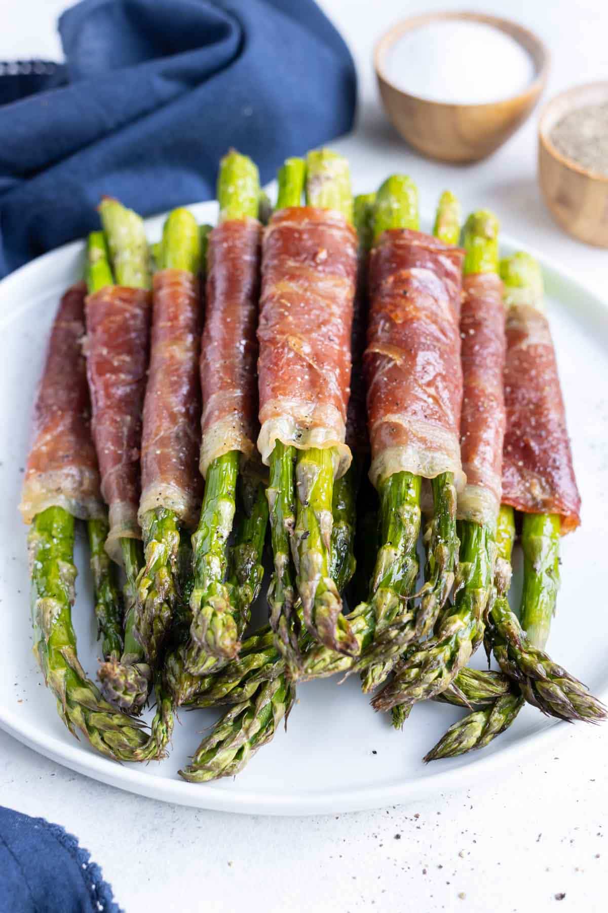 Several asparagus spears individually wrapped in prosciutto and dashed with spices on a white plate. 