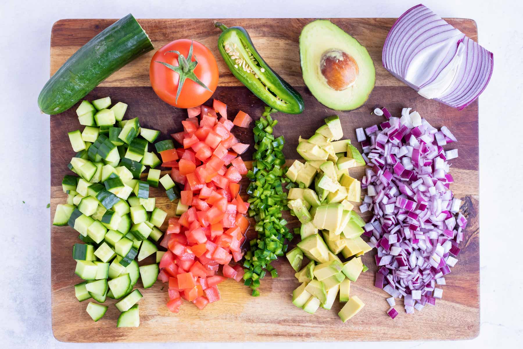 A wooden cutting board with diced cucumber, tomatoes, jalapeno, avocado, and red onion.
