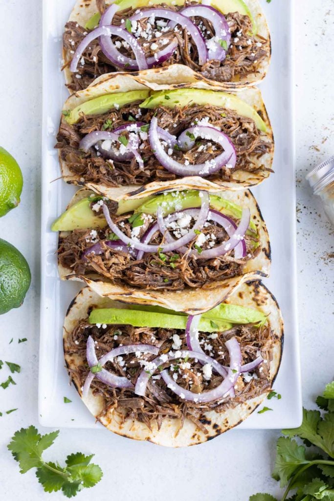 Red onion, cotija cheese, and avocado is placed on top of the barbacoa tacos.