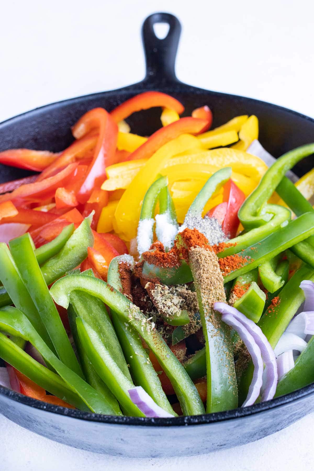 Red, yellow, and green bell pepper slices are cooked in a cast iron skillet with red onions and fajita seasoning.
