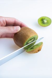 A hand is used to hold the kiwi steady while a knife cuts off the end.