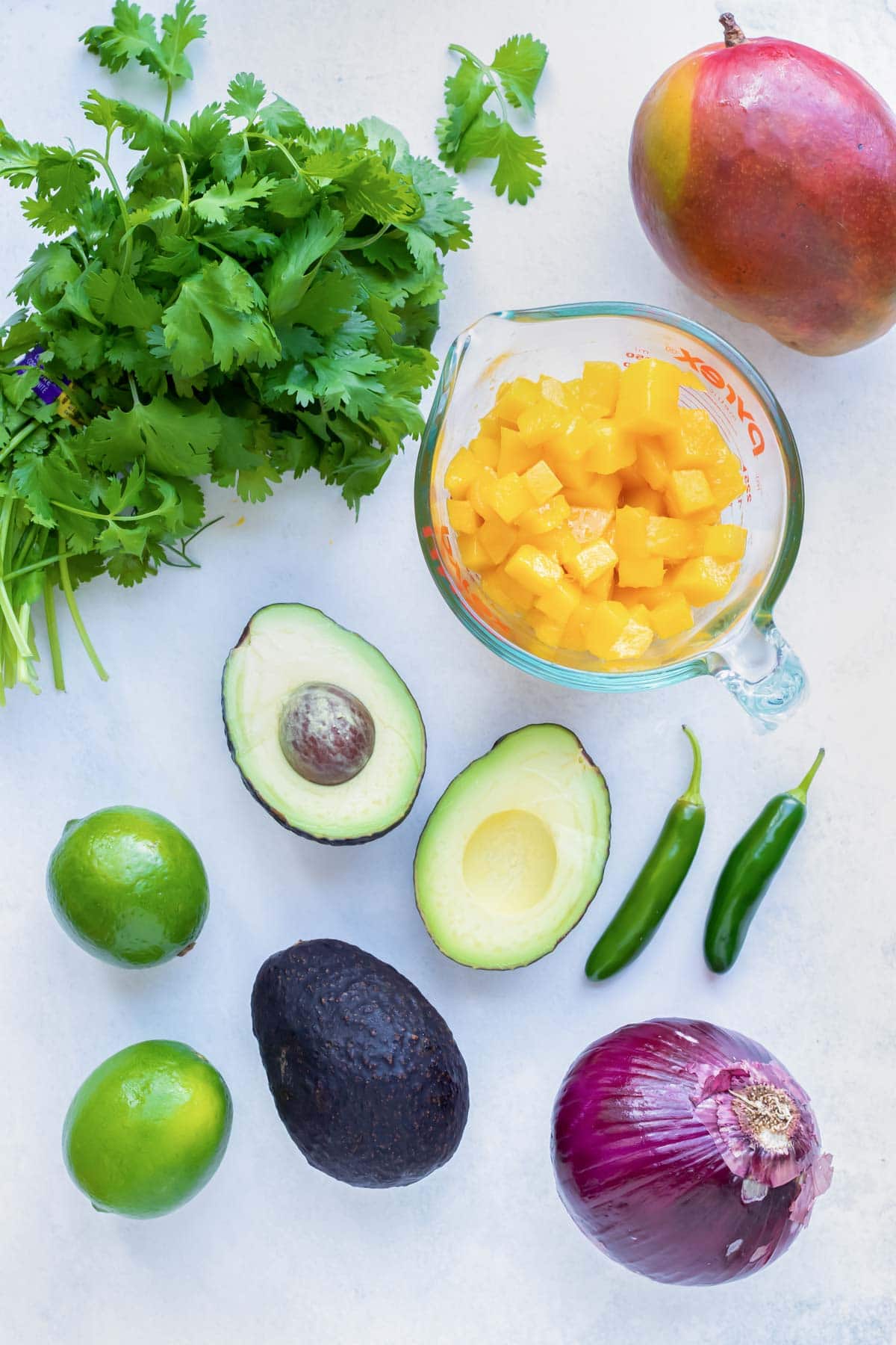 Fresh mango, avocado, serrano peppers, limes, red onion, and cilantro as the ingredients needed for a salsa recipe.
