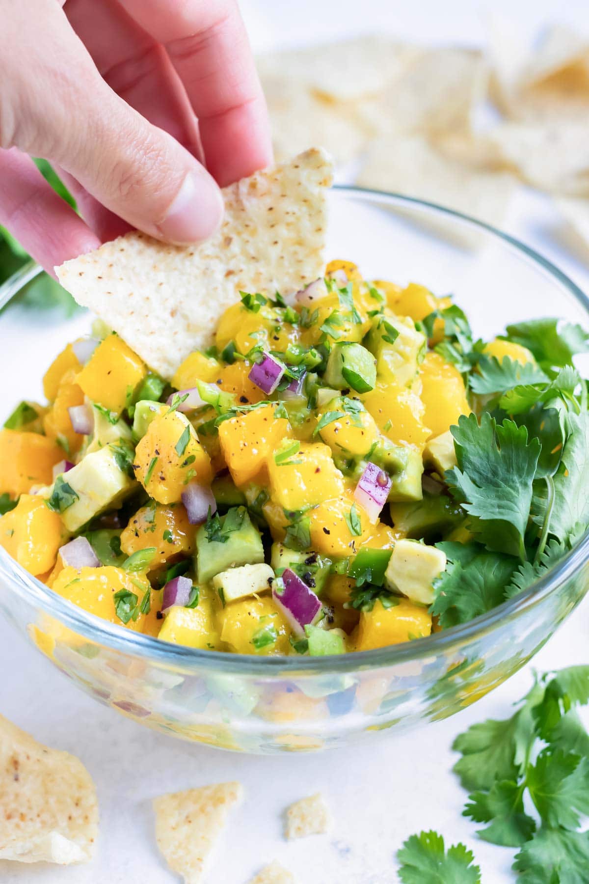 A tortilla chip dipping into a big glass bowl full of mango salsa.