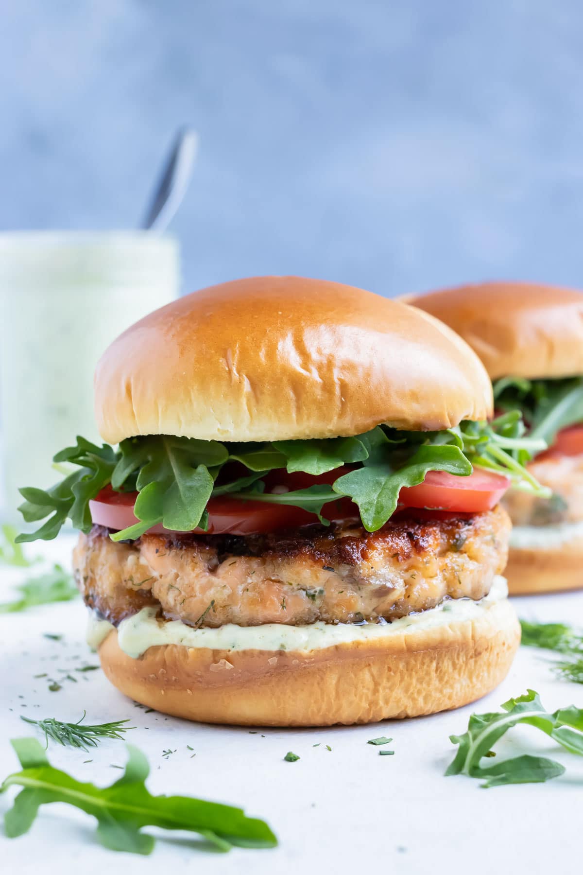 Salmon burgers are served on a bun with tomatoes, lettuce, and avocado sauce.