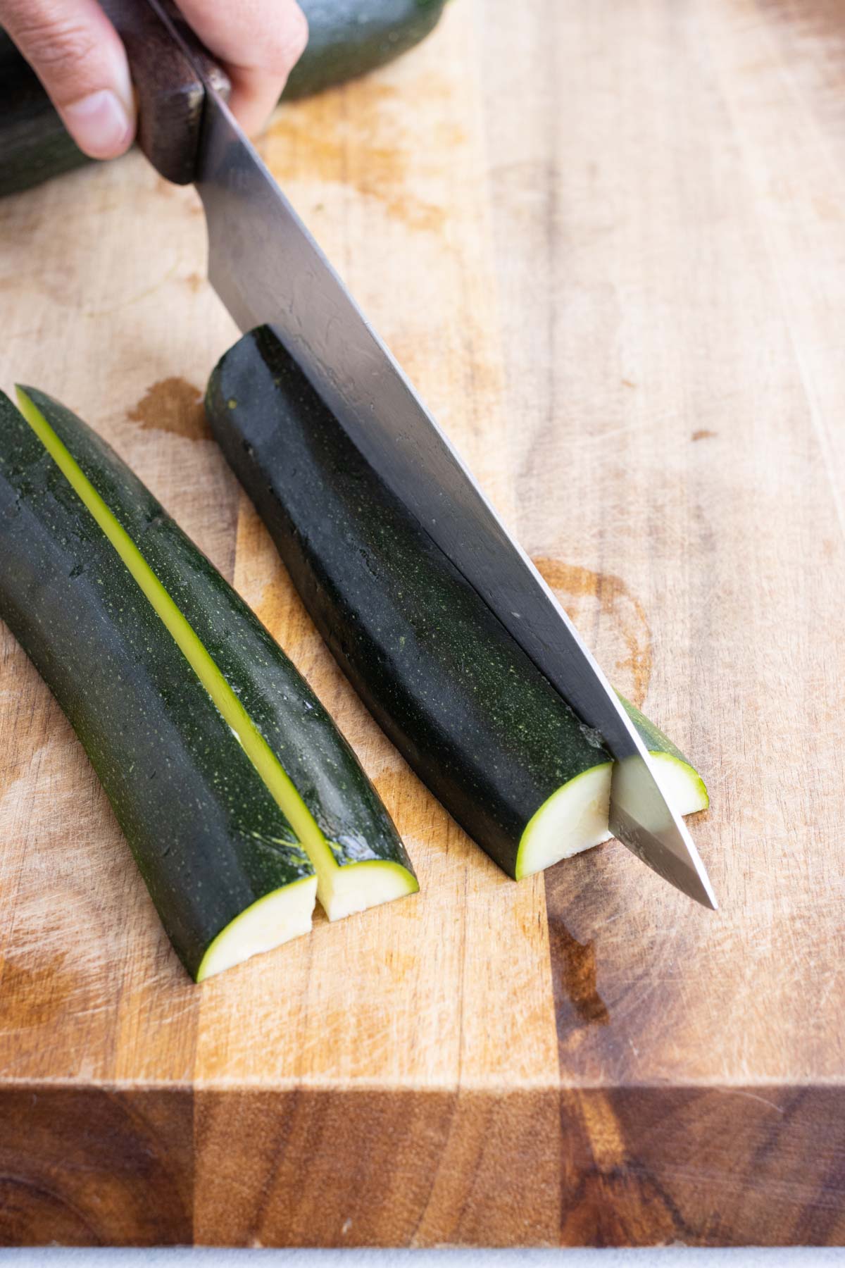 A knife slices a zucchini in quarters on a cutting board.