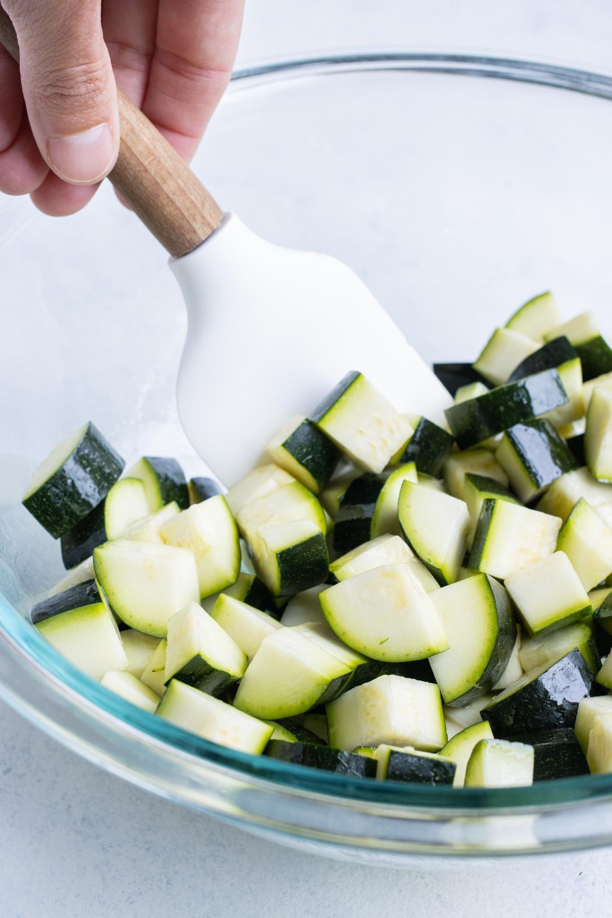 A spatula stirs sliced zucchini in a glass bowl.