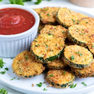 Air fryer zucchini chips are shown on a white plate with dip and fresh parsley.