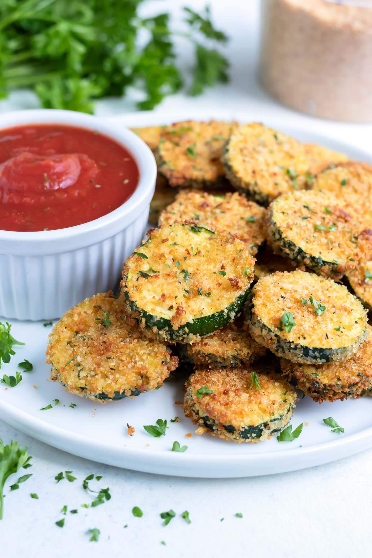 Air fryer zucchini chips are shown on a white plate with dip and fresh parsley.