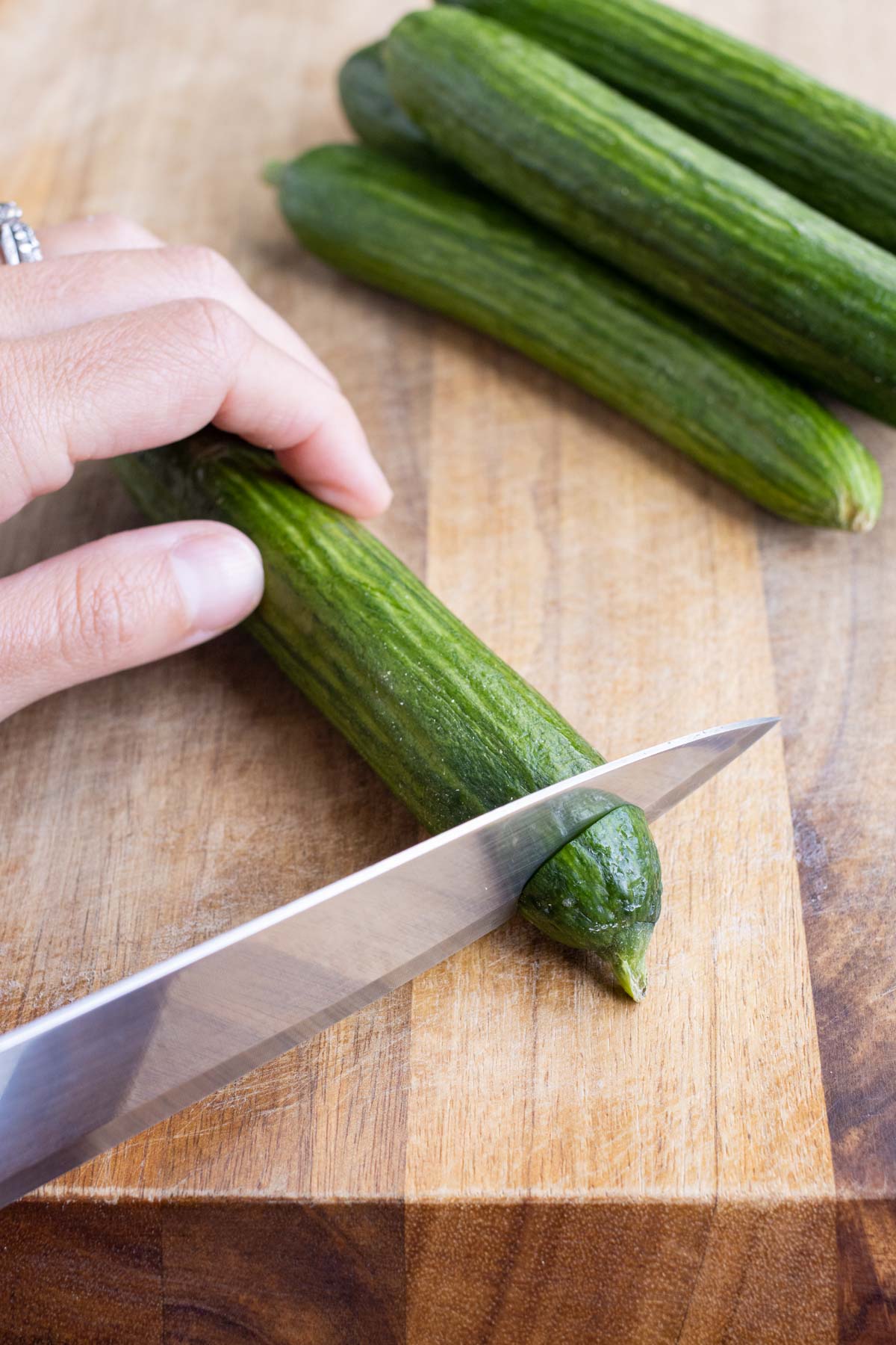 A sharp knife cuts the end off a cucumber on a wooden cutting board.