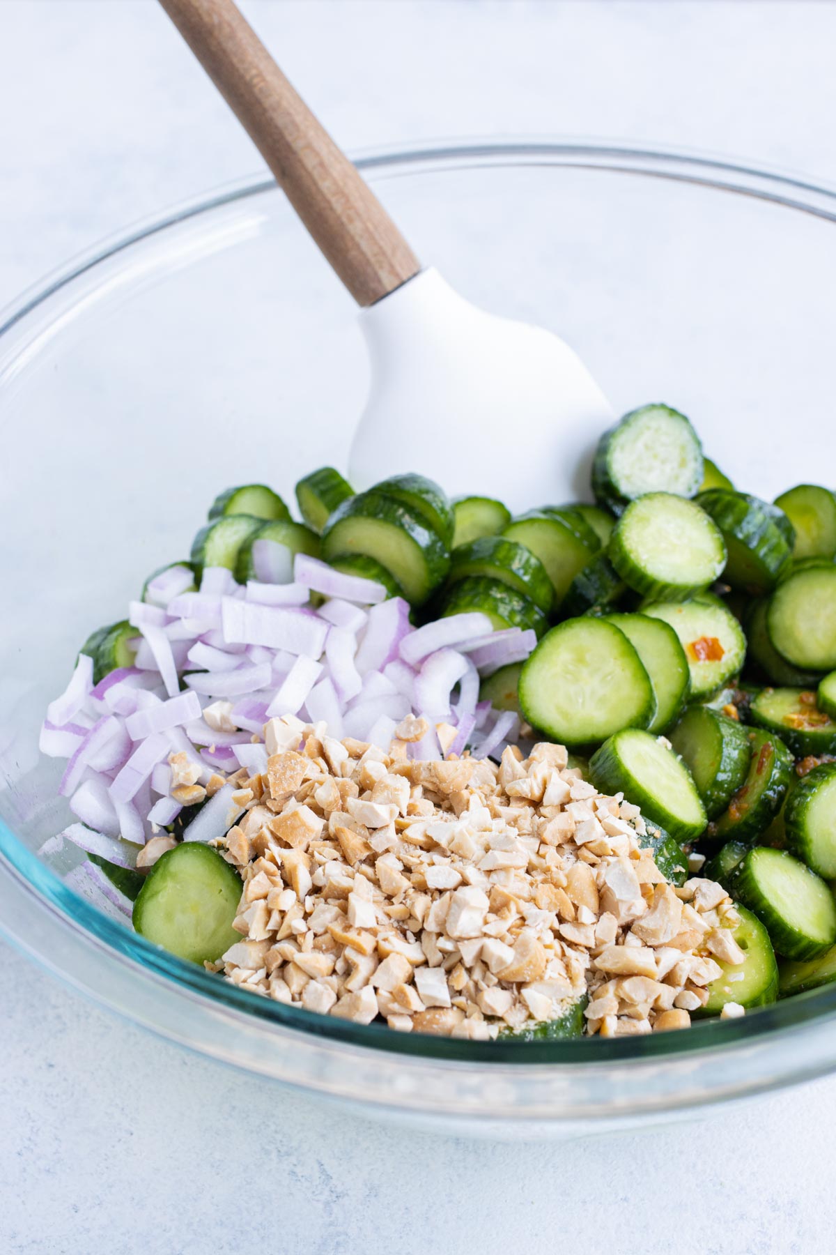 A spatula stirs cashews and onions into cucumbers with dressing on them.