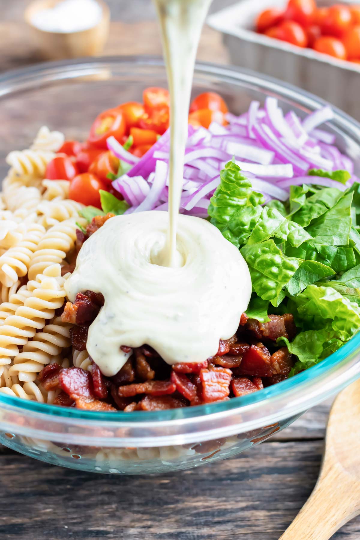 Ranch dressing mix being poured over pasta, bacon, and veggies in a large glass bowl.