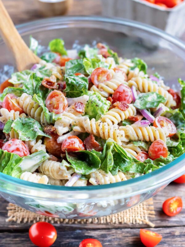 A glass Pyrex bowl full of a BLT pasta salad being mixed together with a wooden spoon.