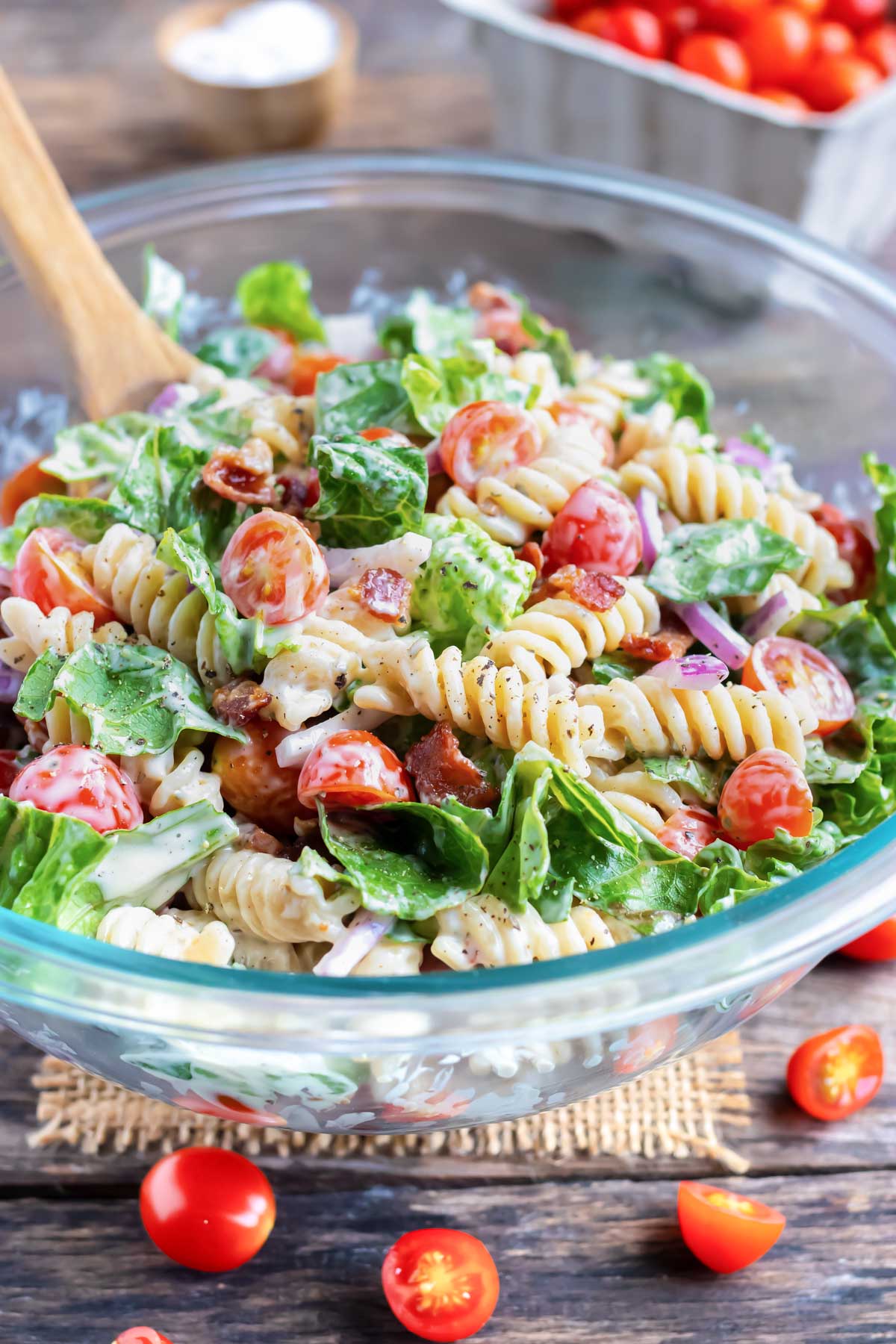 A glass Pyrex bowl full of a BLT pasta salad being mixed together with a wooden spoon.