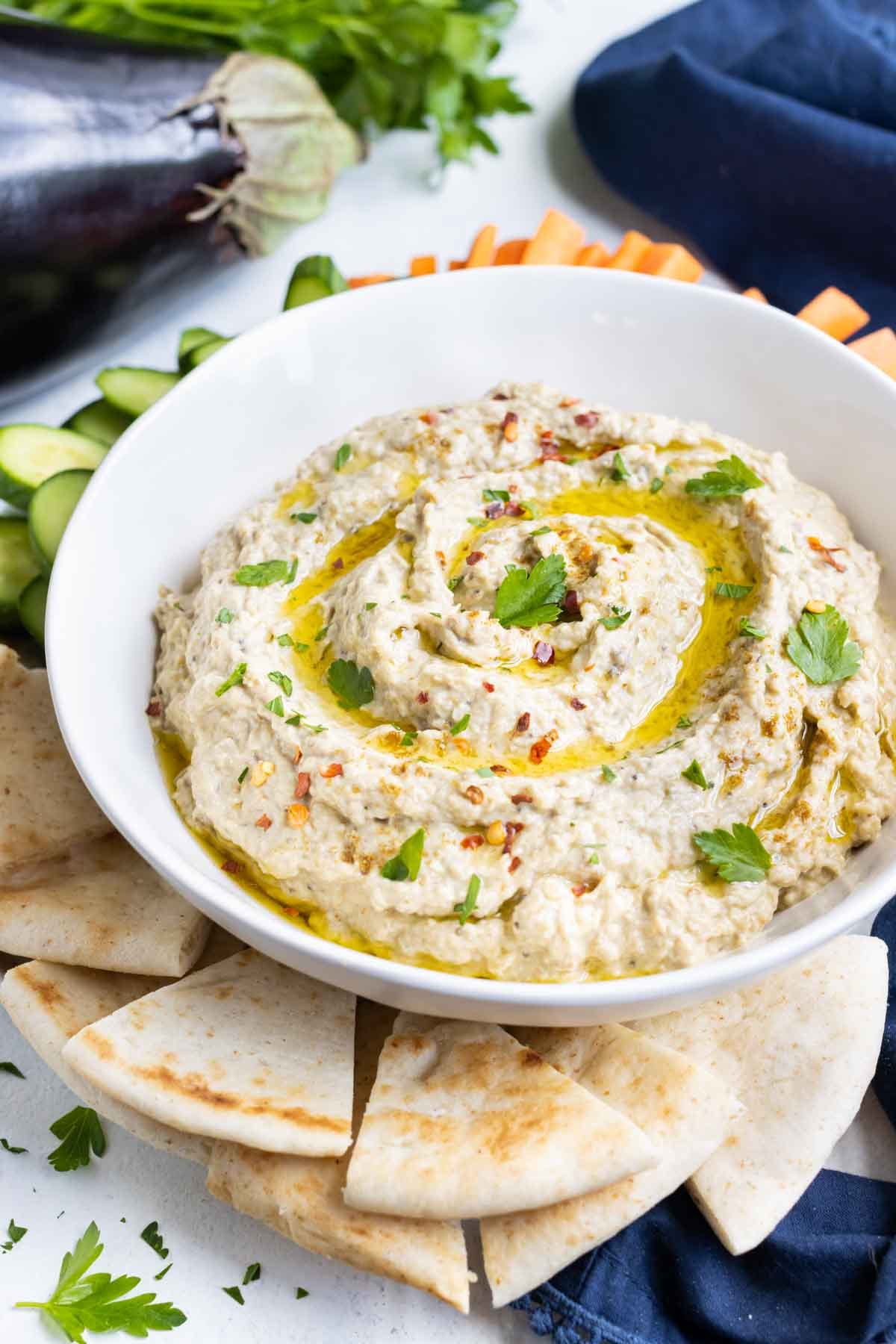 Creamy and healthy eggplant dip in a white bowl surrounded by pita and veggies.