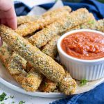 A hand picks up a baked zucchini fry to dip in sauce.