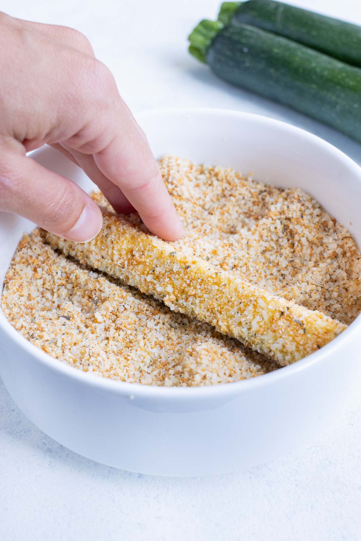 A hand holds a zucchini fry over a bowl with seasonings and breadcrumbs.