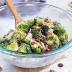 Broccoli Bacon Salad with cranberries in a clear glass bowl with a wooden spoon stirring it.