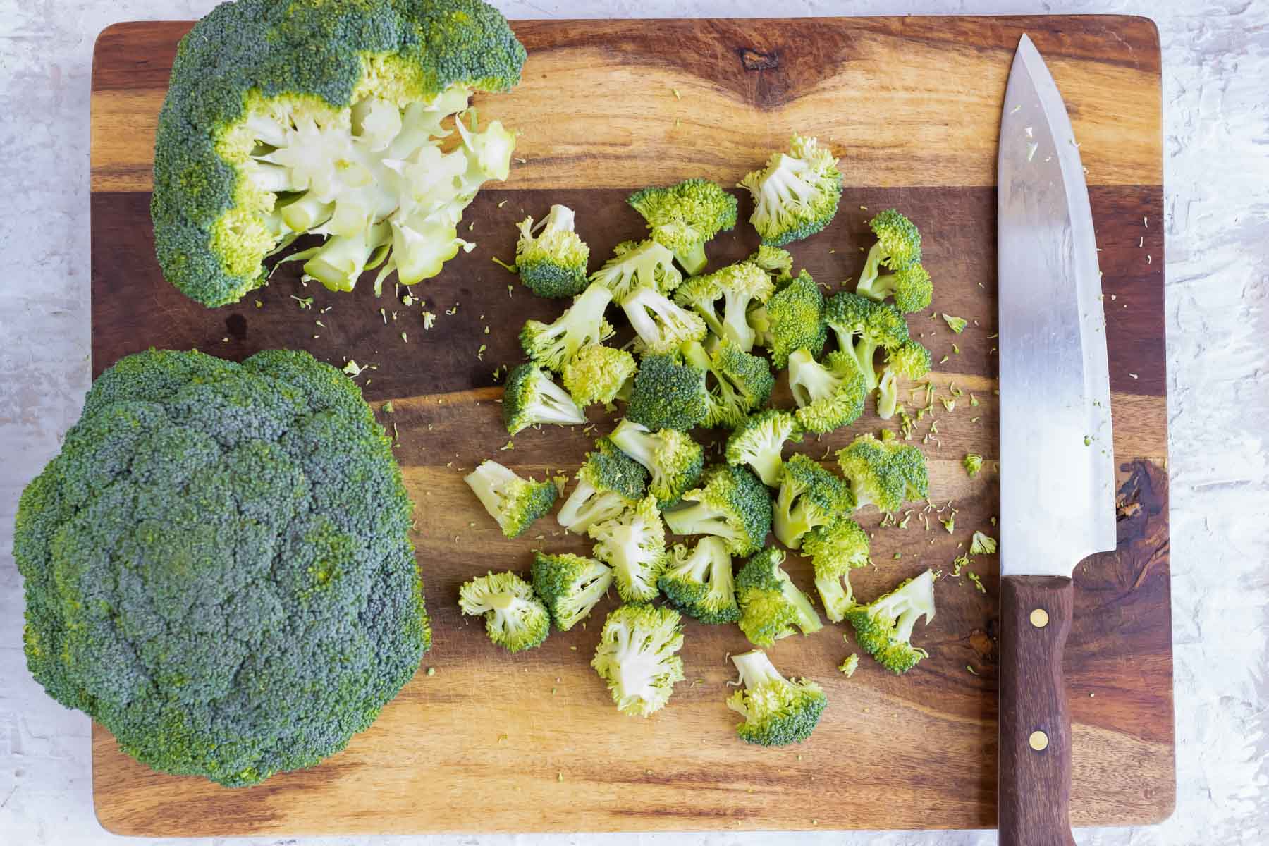 Broccoli florets and stalks chopped with a sharp knife on a wooden cutting board.