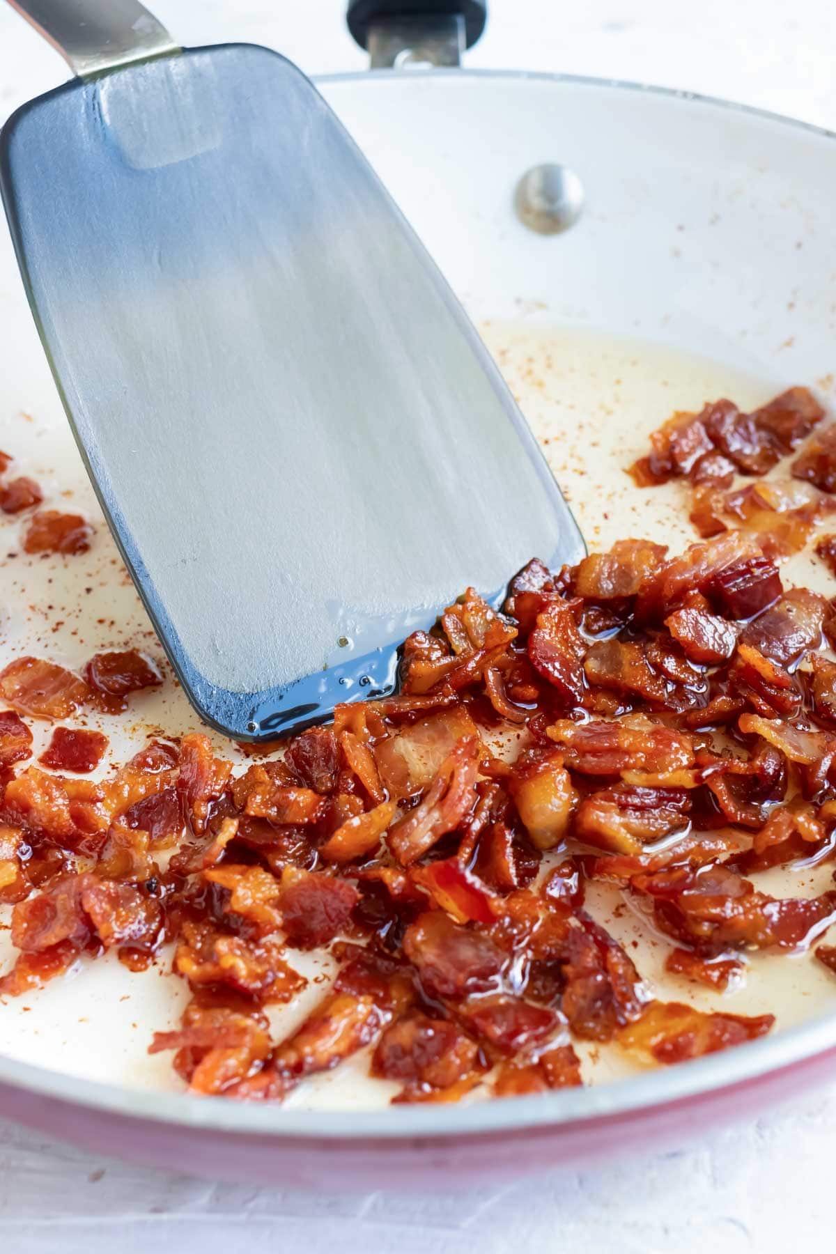 Bacon being cooked in a ceramic skillet with a spatula stirring it.