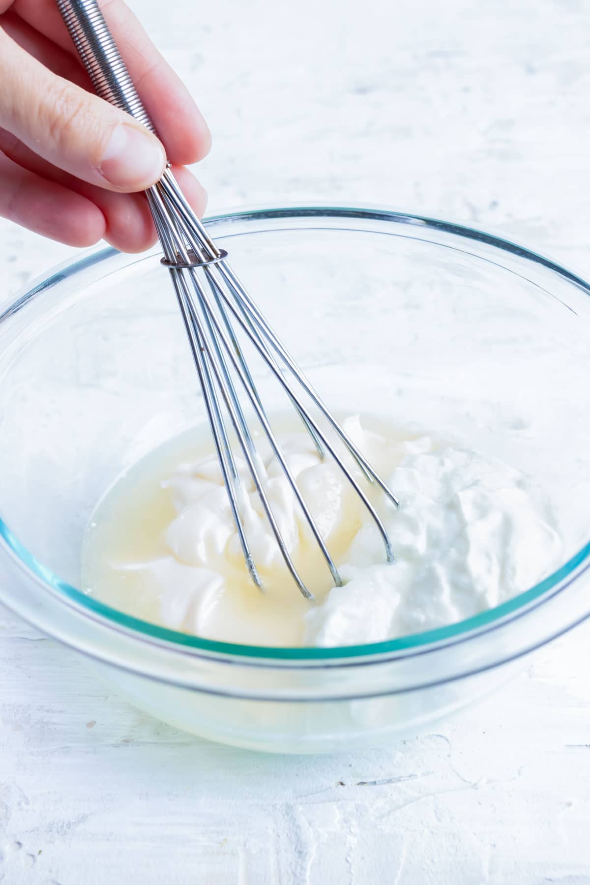 A hand whisking together a broccoli salad dressing made from mayonnaise, white wine vinegar, Greek yogurt, and sugar.