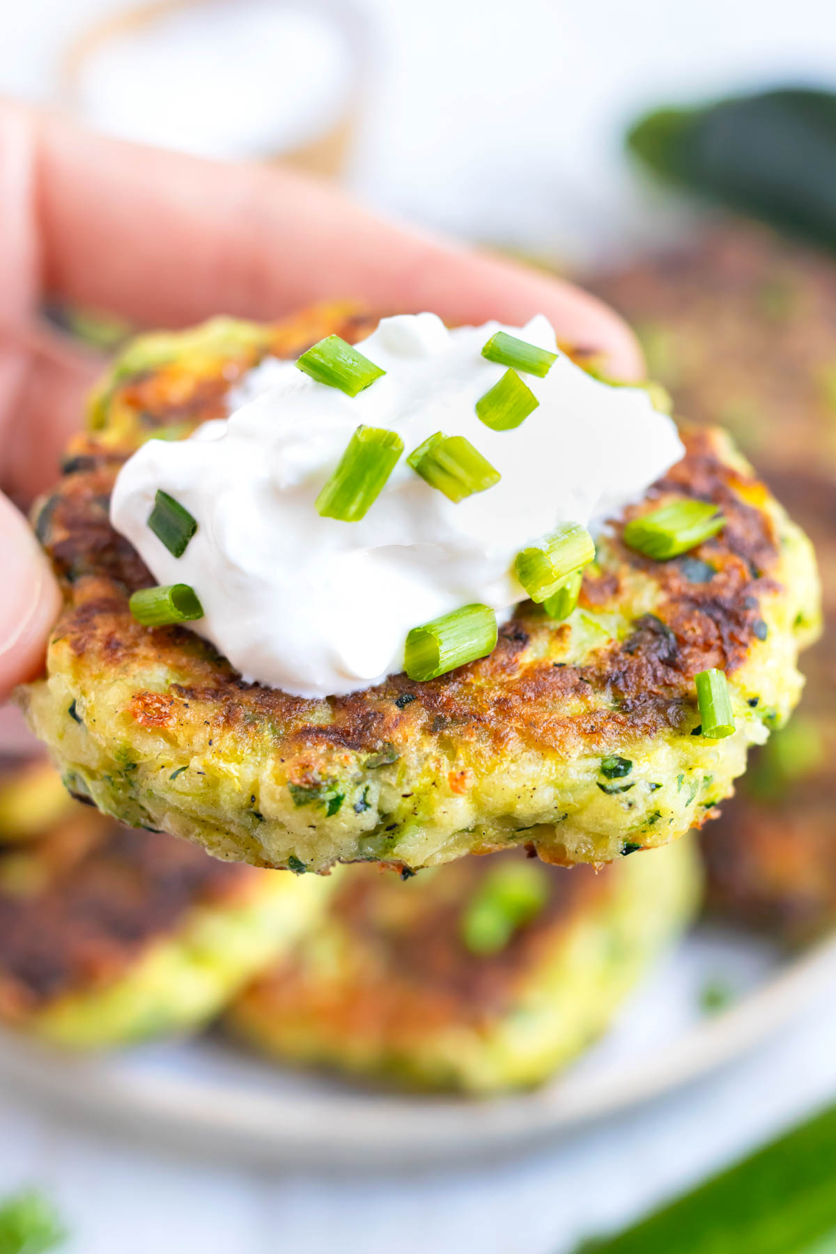 A hand holding a zucchini fritter with sour cream on top and green onions.