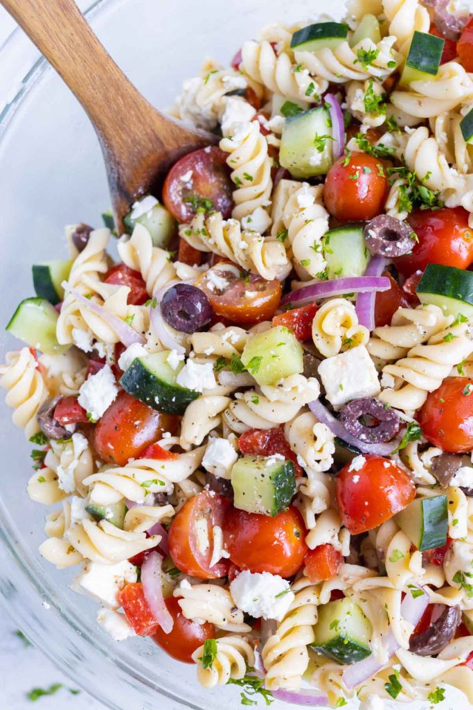 Greek pasta salad is served with a wooden spoon from a bowl.