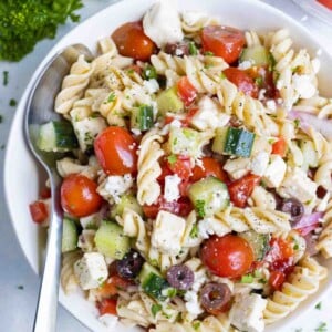 A simple Greek pasta salad is served in a white bowl with a spoon.