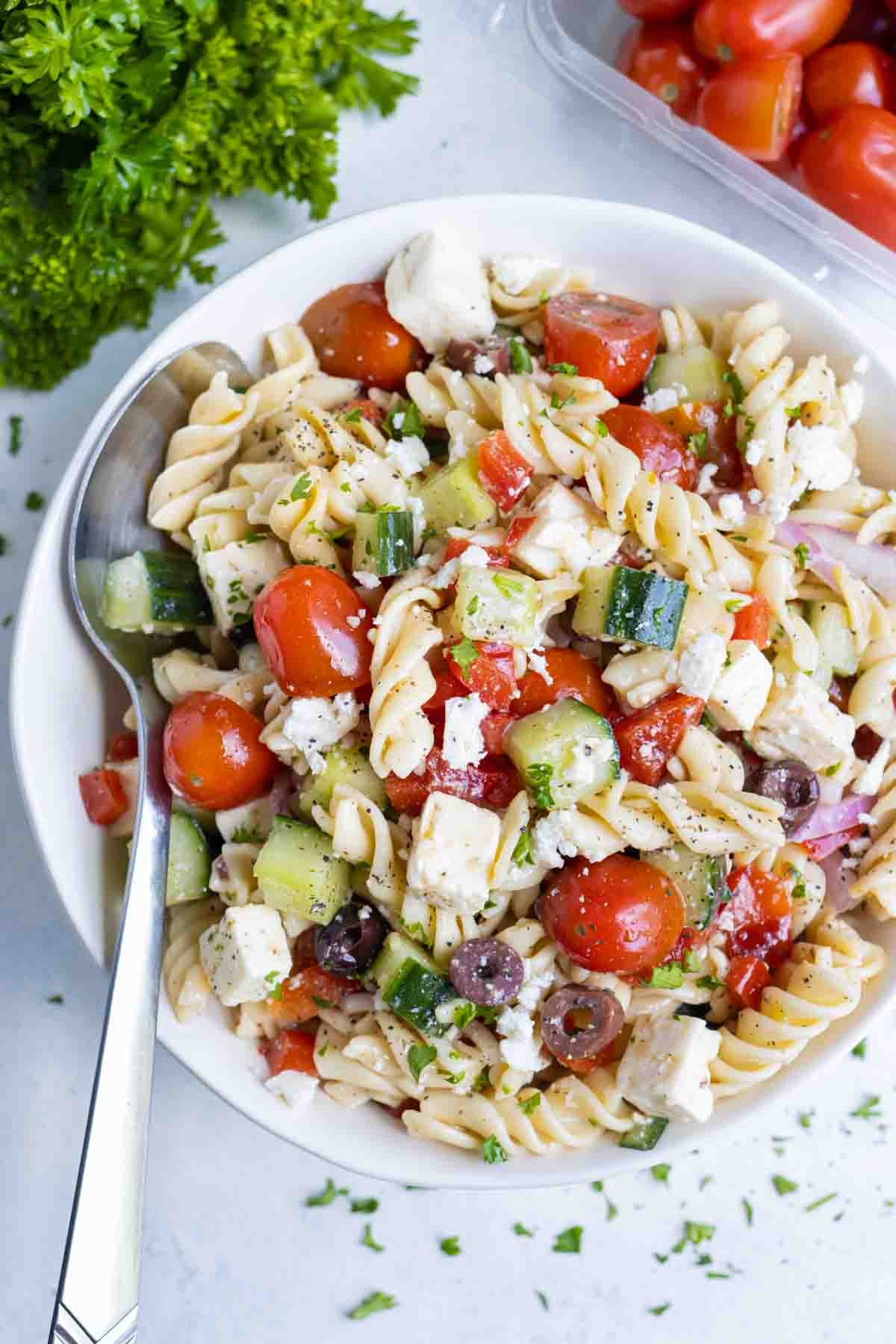 Greek pasta salad is enjoyed from a white bowl with a metal spoon.