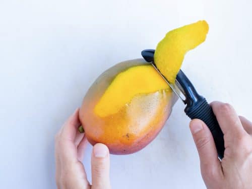 A black vegetable peeler removes the skin from a mango.
