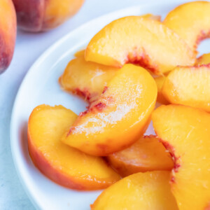 Peeled, sliced, frozen peaches are laying on a white plate on the counter with two whole fresh peaches.