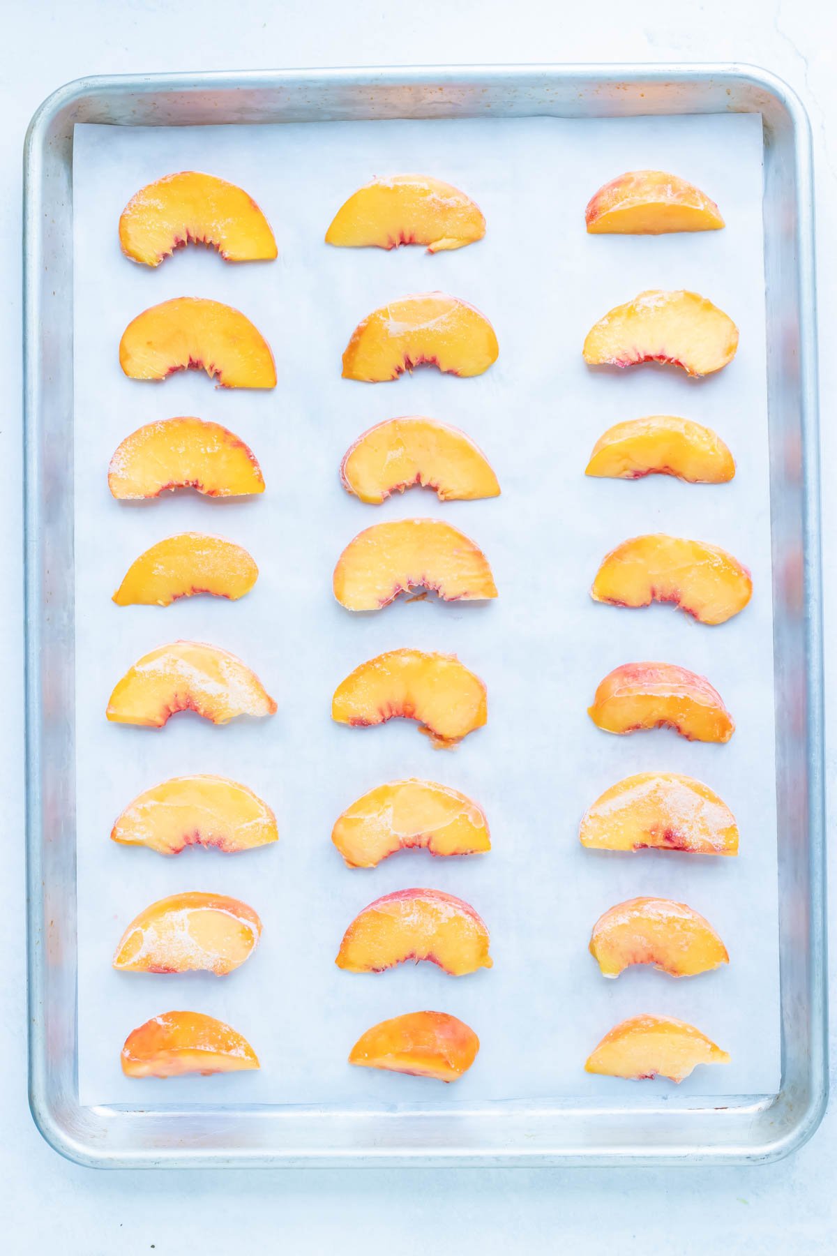 Parchment paper is covering a baking sheet where sliced peached are laid flat to be frozen for future recipes.