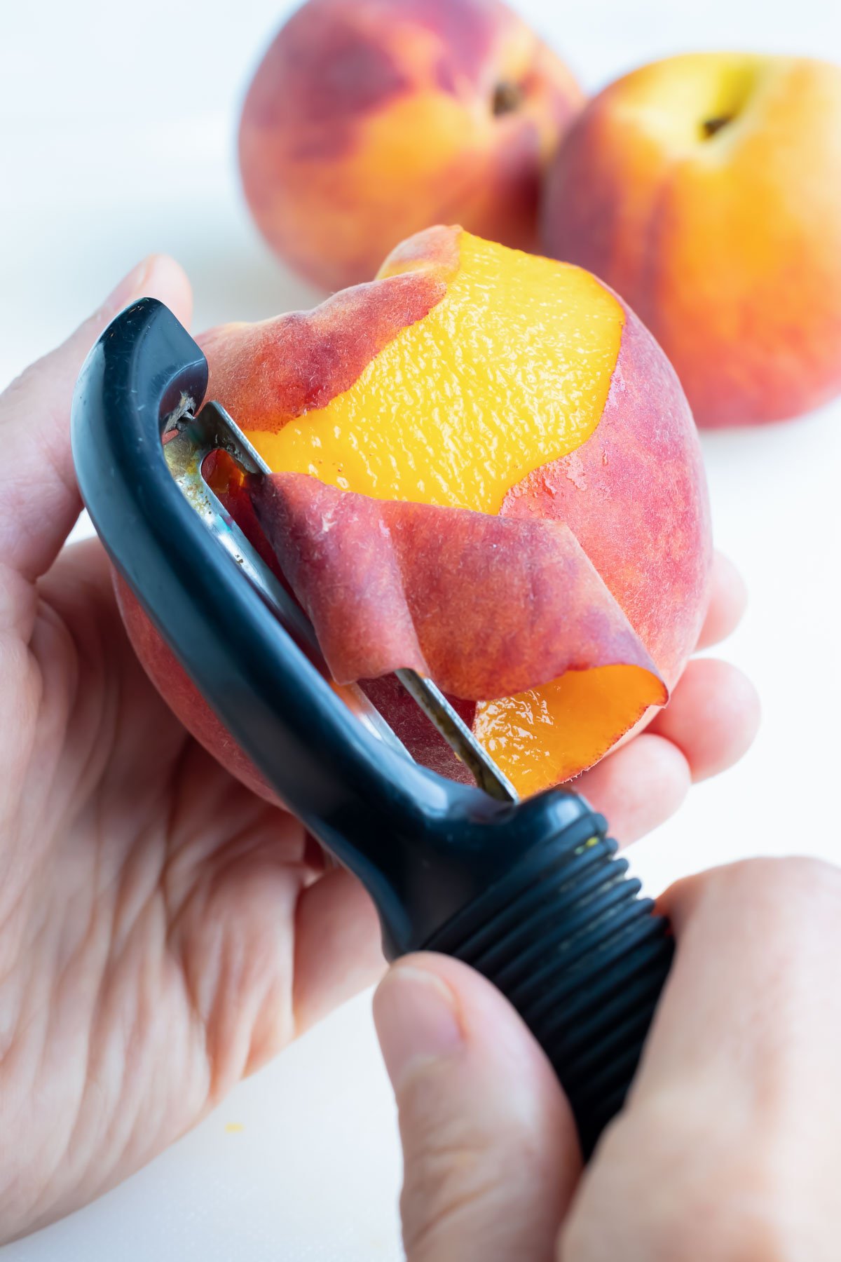 Peeling fruit with a vegetable peeler while two more peaches sit on the counter to be peeled.