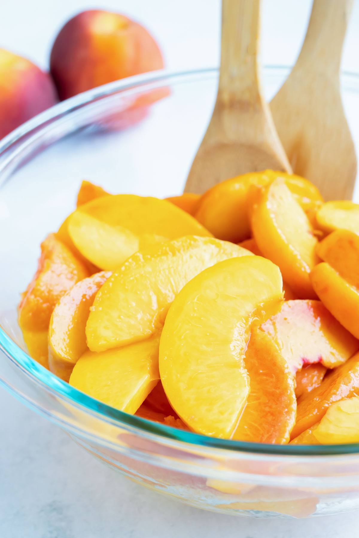 Sliced and peeled peaches are in a glass bowl on the counter with wooden spoons.