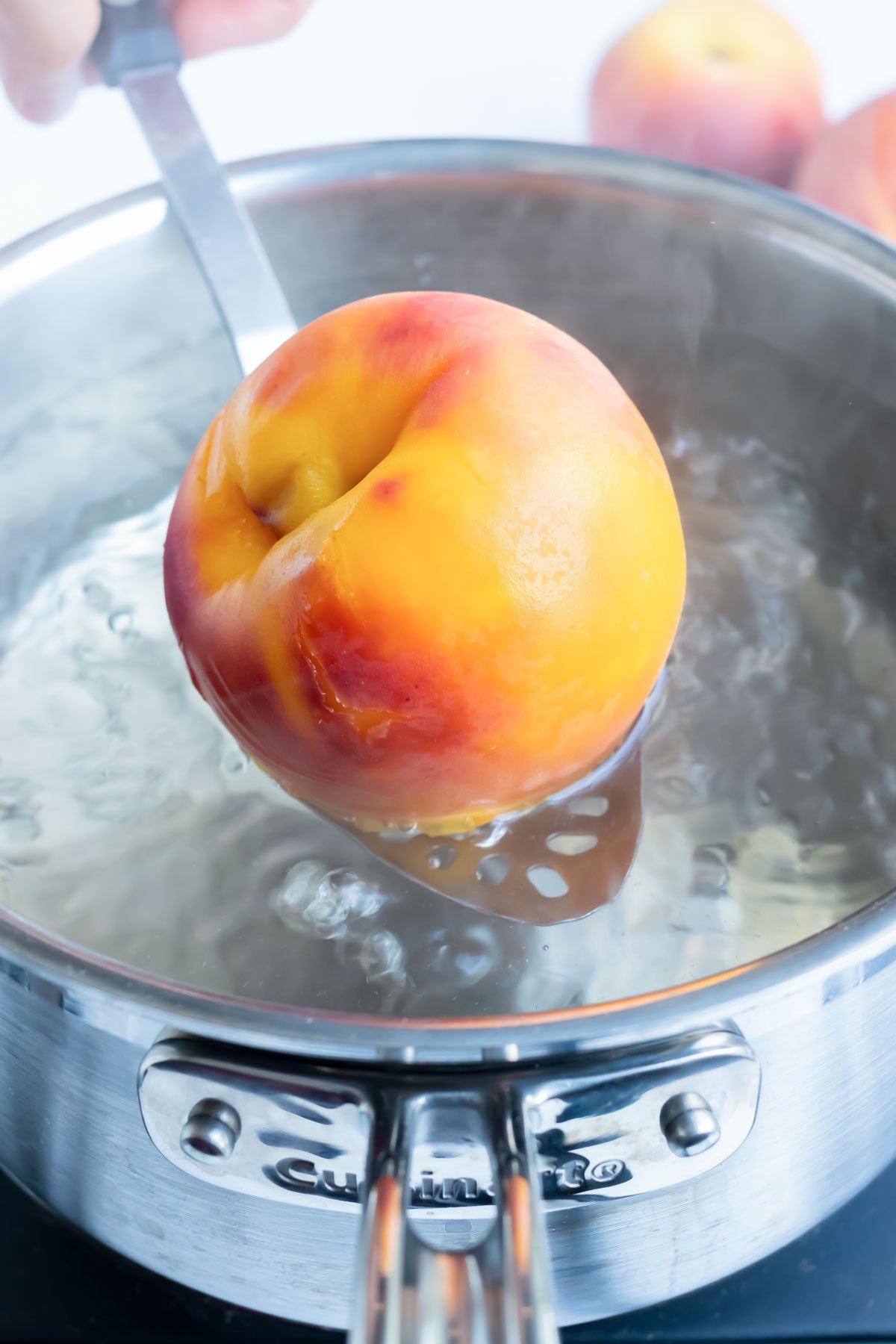 A whole peach is blanched in a pot of hot water and taken out with a metal spoon in preparation for peeling.