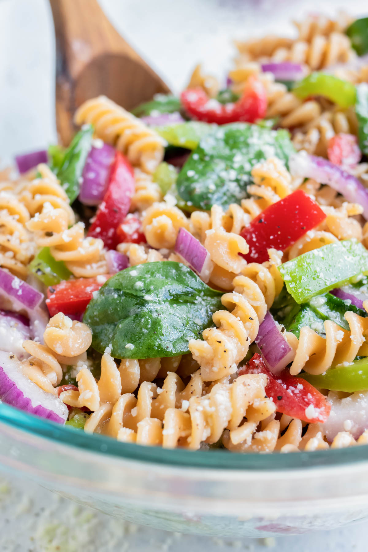 A glass bowl of Italian dressing pasta salad is served from a glass bowl with a wooden spoon.