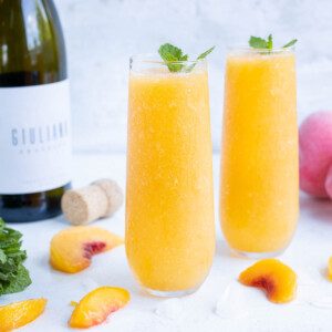 Two flutes with frozen cocktails next to a bottle of prosecco and sliced peaches.