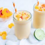 Peach smoothies are placed on the counter with ice, a lime, and a bowl of peaches.