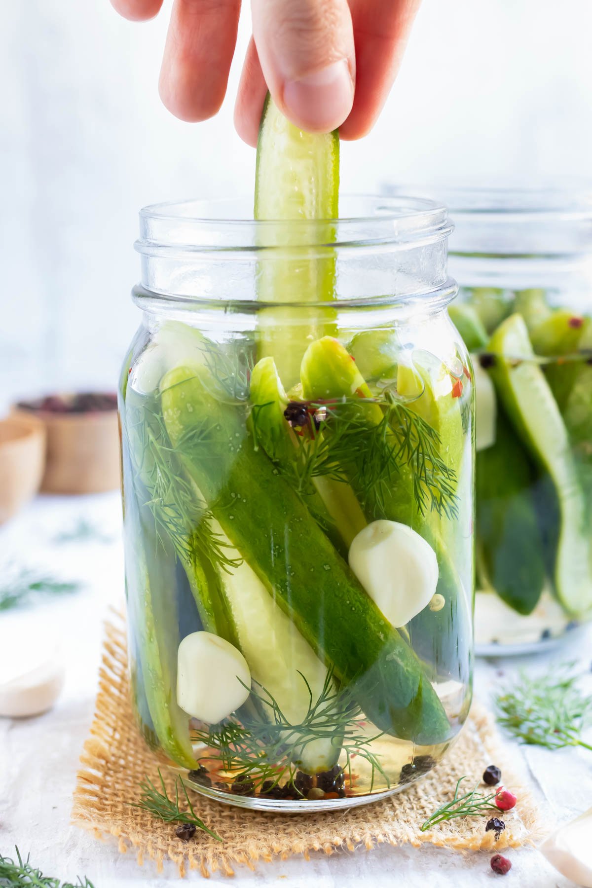 A hand picking up a pickled cucumber spear from a jar of refrigerator dill pickles with another jar in the background.