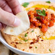 A hand dips some red pepper hummus with a slice of pita bread.