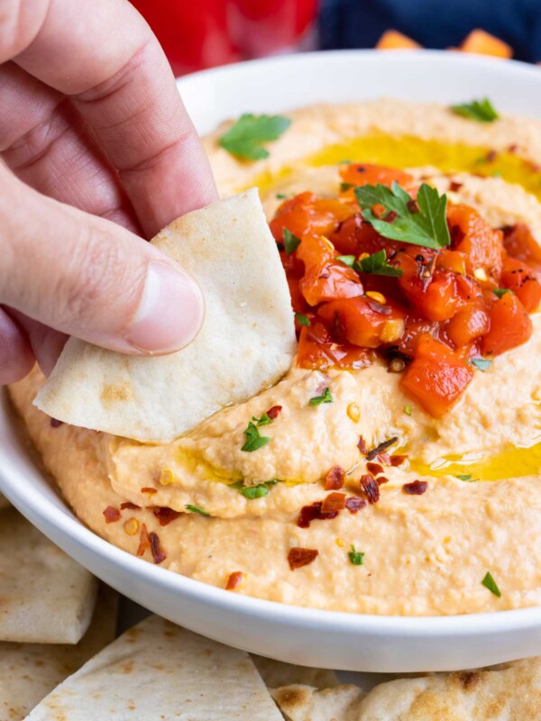 A hand dips some red pepper hummus with a slice of pita bread.