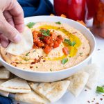 A piece of pita dips some roasted pepper hummus out of a bowl.