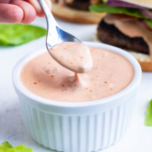 Homemade secret sauce is being scooped up from a white bowl with a spoon to put on a burger.