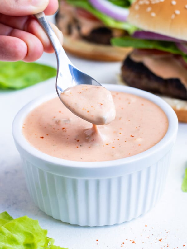 Homemade secret sauce is being scooped up from a white bowl with a spoon to put on a burger.