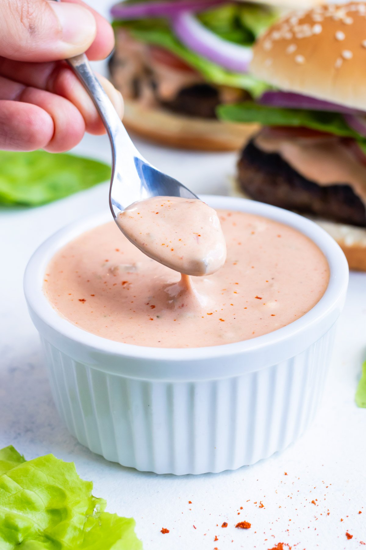 Homemade secret sauce is being scooped up from a white bowl with a spoon to put on a burger.