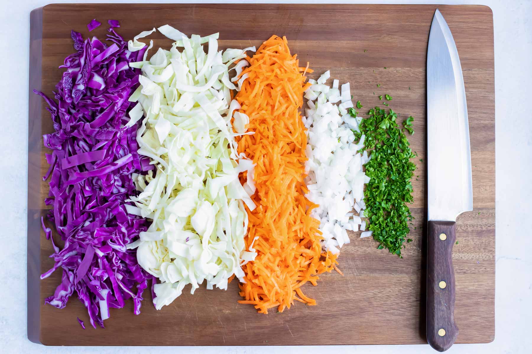 Shredded red cabbage, green cabbage, carrots, onion, and parsley on a chopping board.
