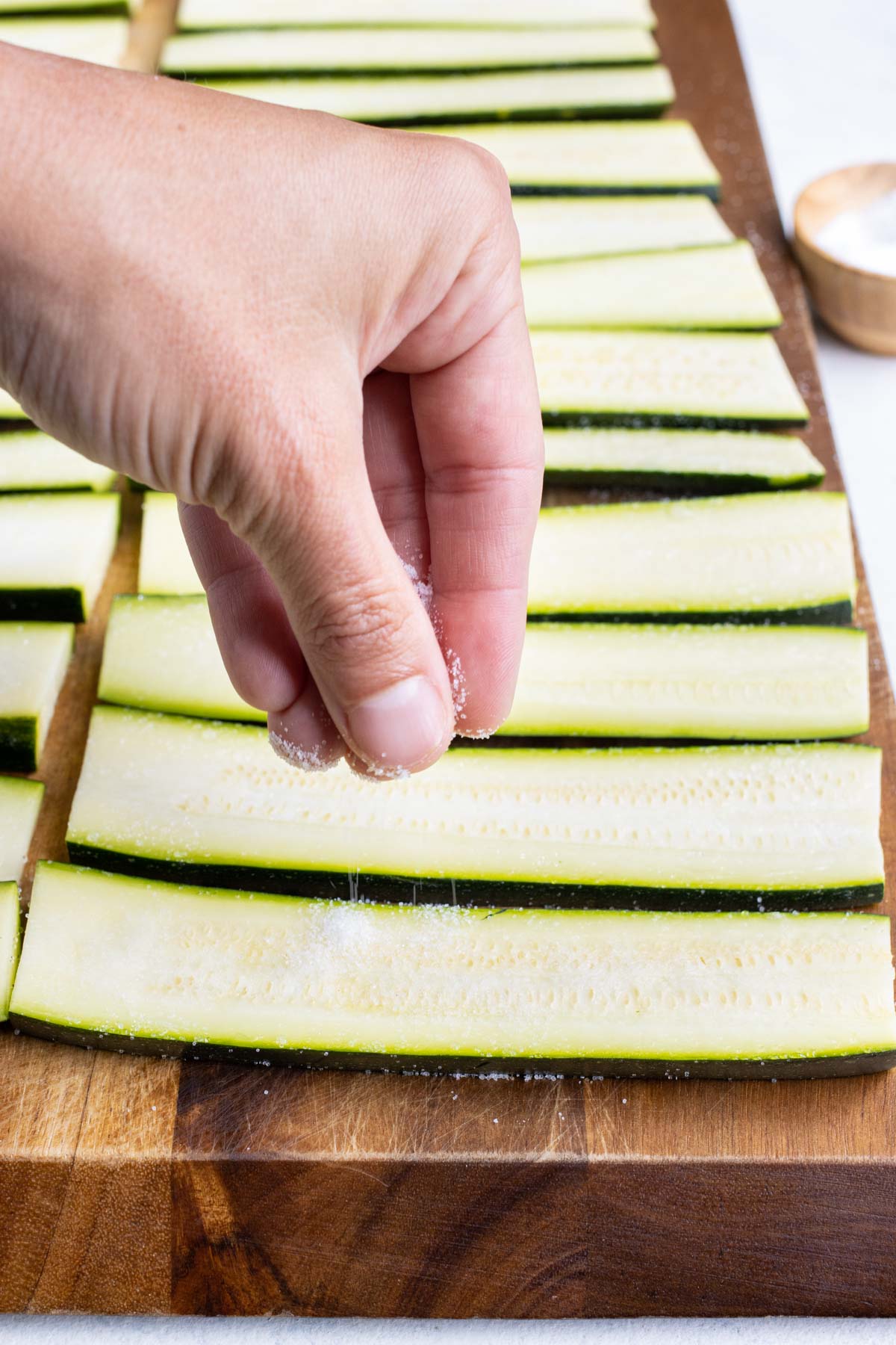 A hand sprinkles salt onto zucchini strips.