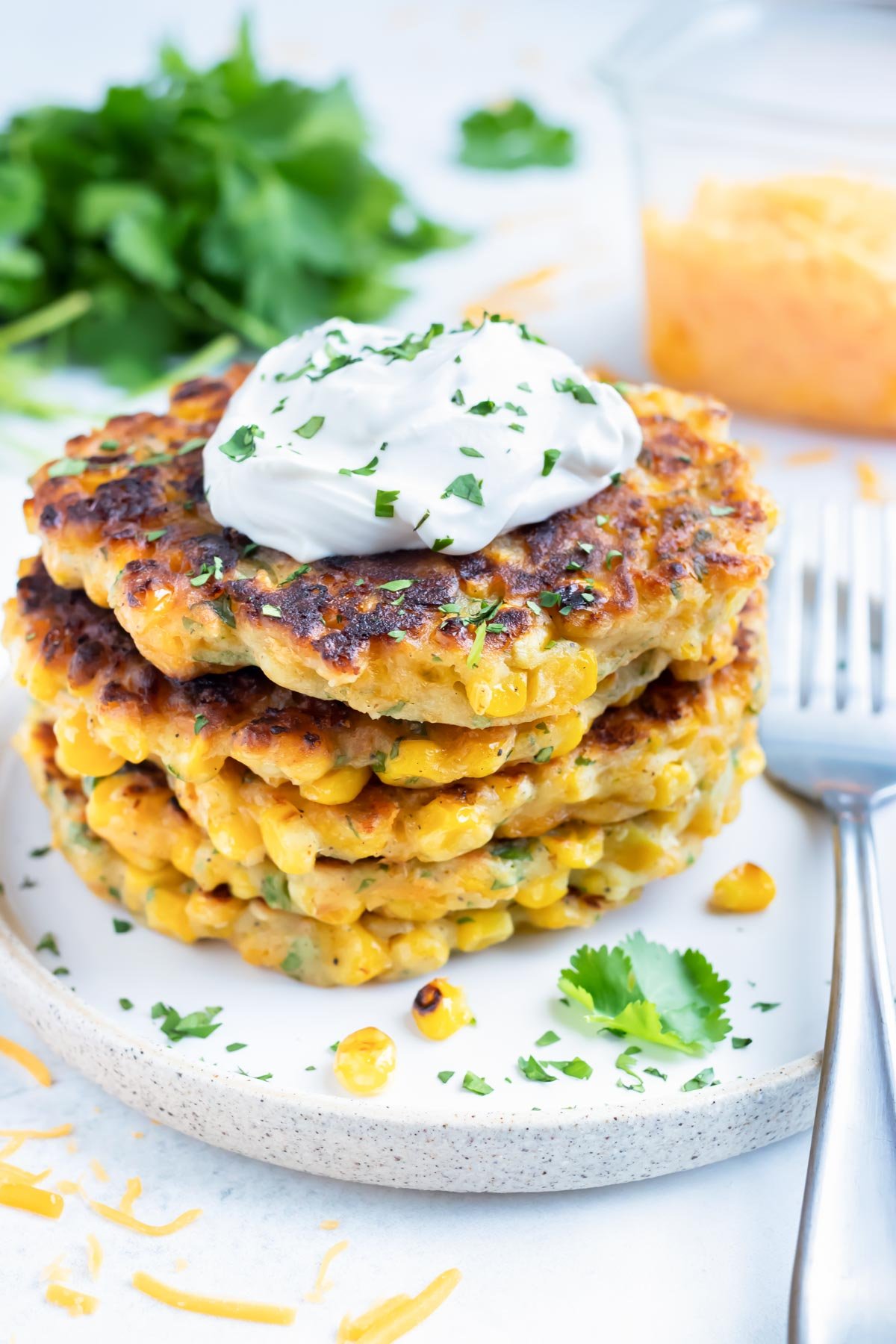 Southern Fried Corn Fritters with a tub of shredded cheddar cheese and cilantro behind them.