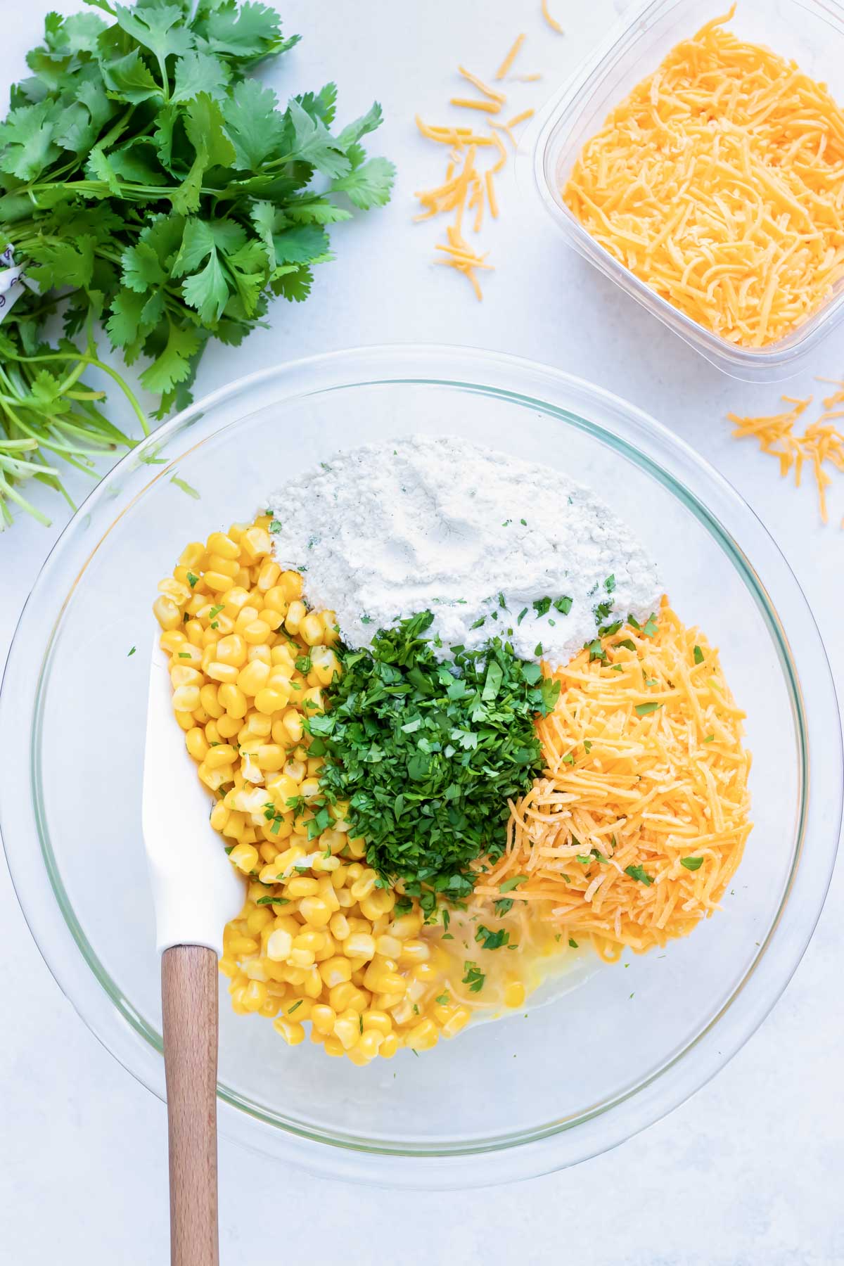 A clear glass mixing bowl with corn kernels, flour, shredded cheddar cheese, and cilantro.