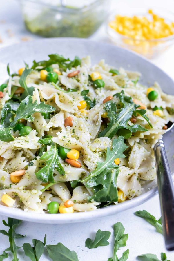Cold pesto pasta salad is served from a white bowl with a metal spoon.