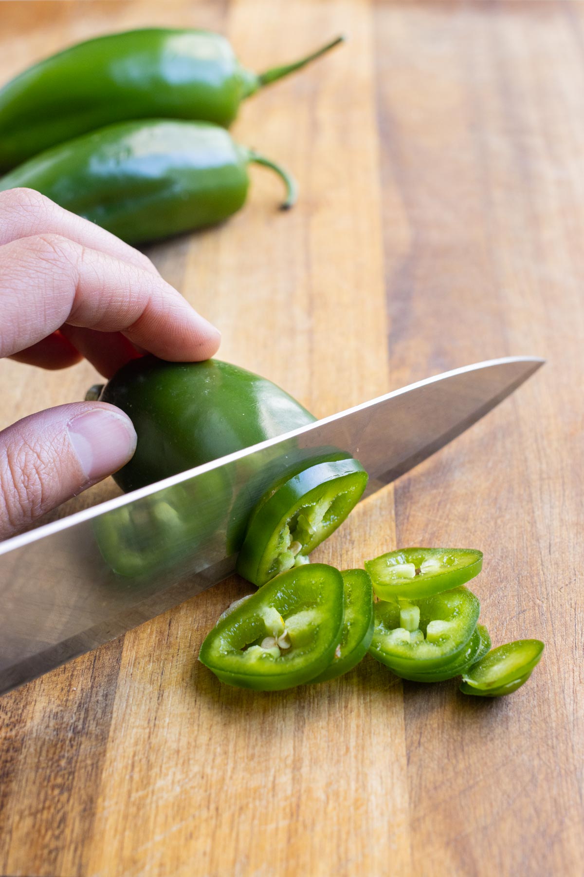 A sharp knife slices a jalapeño on a wooden cutting board.